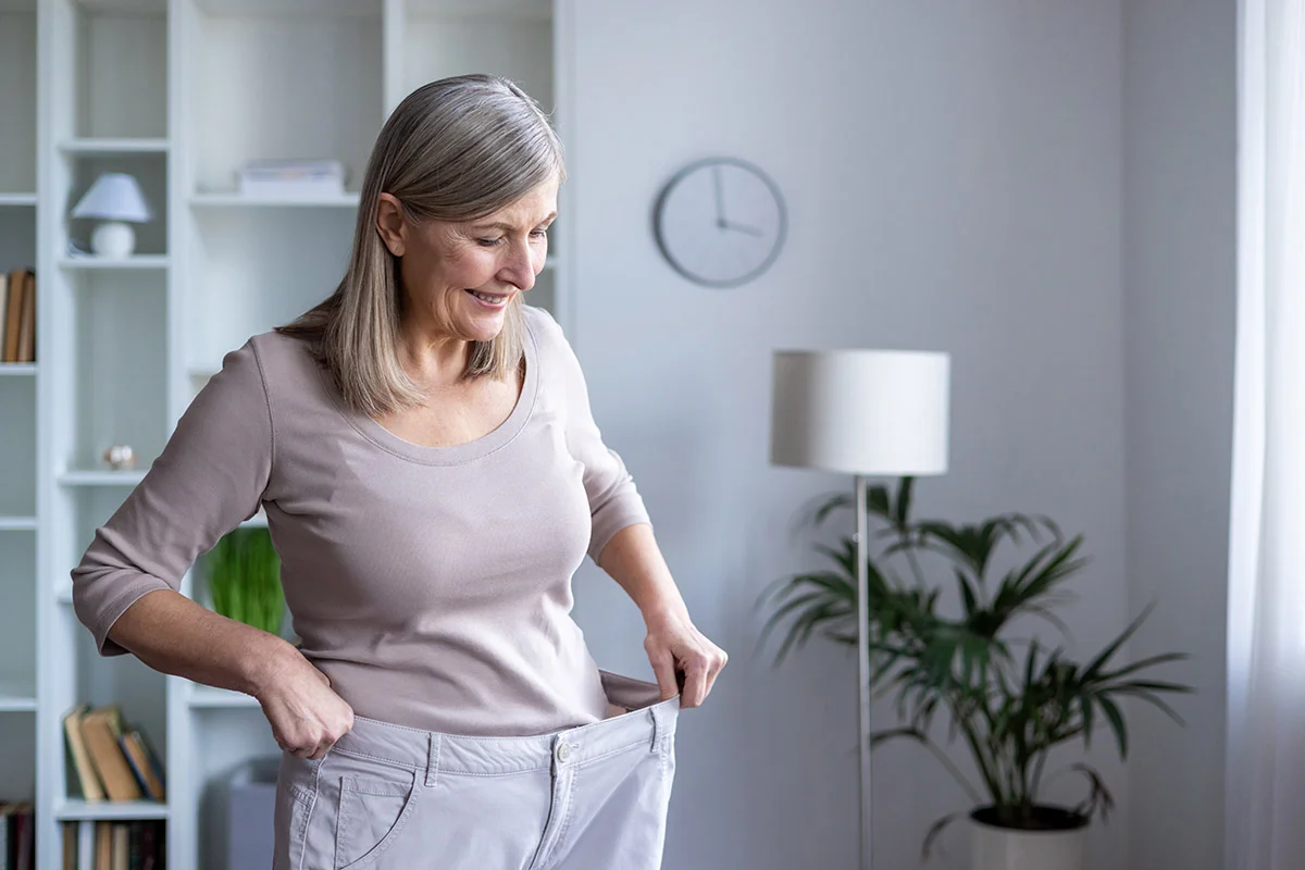 senior woman smiling holding the waist of a pair of oversized pants showing her total weight loss